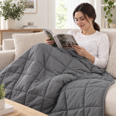 Woman reading on a sofa with the gray weighted throw blanket over her lap and legs